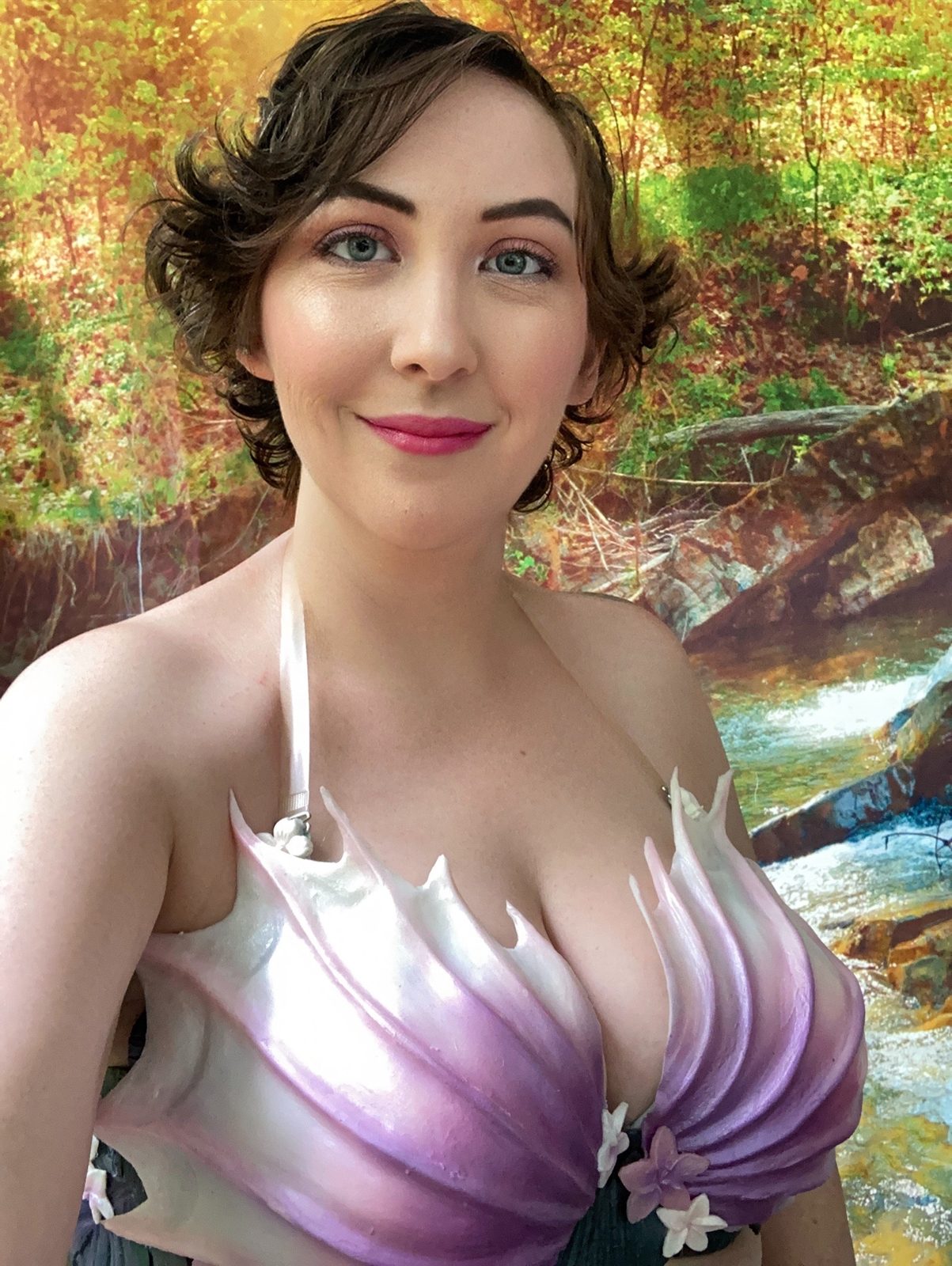 The storyteller mermaid in her shell top, smiling by a forest stream